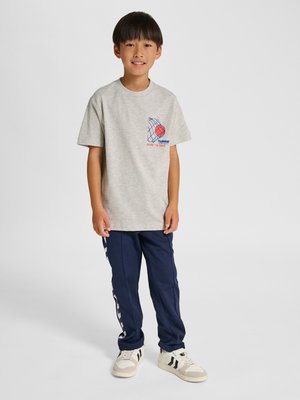 En ung dreng står iført en lysegrå T-shirt med et rødt basketball- og netgrafik, marineblå træningsbukser og hvide sneakers med sorte striber.