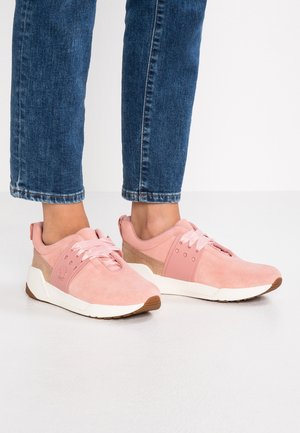 Voeten in roze suède sneakers met witte zolen en satijnen veters, gecombineerd met een blauwe spijkerbroek tegen een effen witte achtergrond.