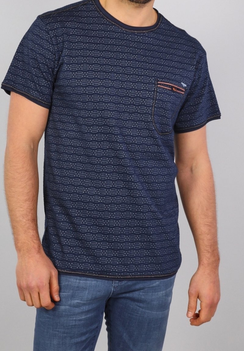 Homme portant un t-shirt à manches courtes à motifs bleu marine avec une poche poitrine et un jean bleu, se tenant devant un fond uni.