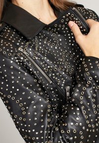 Giacca in pelle nera con dettagli in metallo, colletto e tasca con zip. Superficie texture con accenti circolari distribuiti uniformemente.