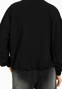 Czarna kurtka przeciwdeszczowa o luźnym kroju, z elastycznymi mankietami i zaokrąglonym dołem. Materiał jest gładki i lekki, z minimalnymi zdobieniami.