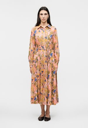 Γυναίκα στέκεται φορώντας ένα midi φόρεμα με μακριά μανίκια και γιακά, με πορτοκαλί, ροζ, μπλε και πράσινο λουλουδάτο μοτίβο, και μαύρα επίπεδα παπούτσια.