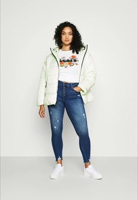 Doudoune vert clair avec des bordures noires, t-shirt graphique blanc avec un motif floral, jeans skinny bleus déchirés et baskets blanches.