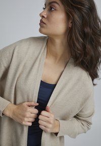 Beige Cardigan aus weichem Strickmaterial, offene Front, lange Ärmel, gerippte Bündchen, über einem marineblauen Tanktop getragen. Sichtbares Detail des strukturierten Stoffes.