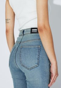 Primer plano de una persona que lleva vaqueros de cintura alta de color azul claro con una etiqueta negra de DRDENIM en la pretina y una camiseta sin mangas blanca.