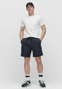 Vit T-shirt med kort ärm och ficka, kombinerad med marinblå shorts. Grå träningsskor och svart keps fulländar outfiten, stående mot en neutral bakgrund.
