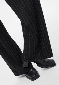 Musta raitakuosinen, leveälahkeiset housut, yhdistettyinä mustiin, neliömäisillä kärjillä varustettuihin nilkkureihin valkoisella taustalla.