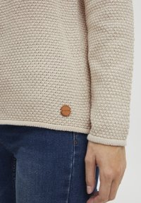 Pull tricoté beige clair avec un motif tissé texturé et un logo en bois au bas, associé à un jean en denim bleu foncé.