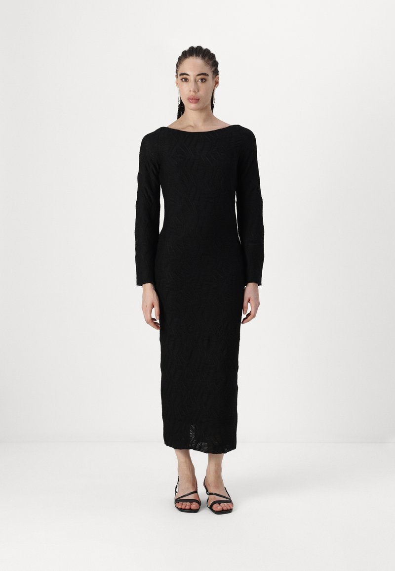 Vestido preto de mangas longas com padrão texturizado geométrico, silhueta ajustada, decote barco e comprimento até a metade da panturrilha, combinado com sandálias pretas de tiras.