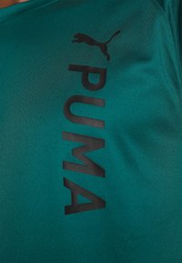 Tyrkysové sportovní tričko s matně černým logem "PUMA" a siluetou pumy na levém horním rohu. Hladká, mírně lesklá textura.