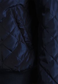 Vadderad marinblått tyg med en glänsande yta, med en sidoficka och en ribbad stickad nederkant.