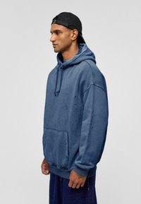 Blauw katoenen pullover hoodie met een kangoeroezak, een capuchon met trekkoord en versleten details, gedragen met donkere blauwe broek.