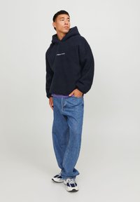 Navy fleece hoodie with a relaxed fit and front pocket, paired with loose blue denim jeans and white and navy sneakers.