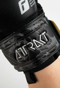 Supporto per polso nero con design testurizzato, cinturino regolabile e logo riflettente "ATRAKT" su una base opaca con motivi sottili.