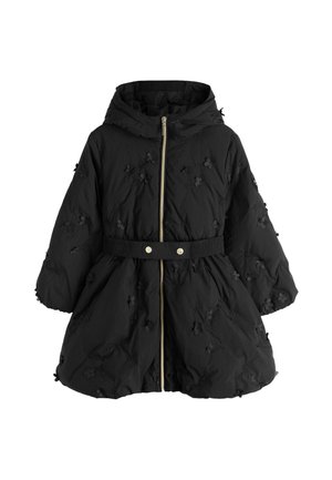 Girl with long wavy hair wearing a belted black puffer coat, black pants, and white sneakers standing on a gray floor against a light wall.