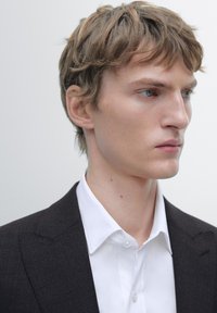 Giacca da completo nera con trama sopra una camicia bianca abbottonata. Il soggetto ha capelli corti castano chiaro ed espressione facciale neutra.