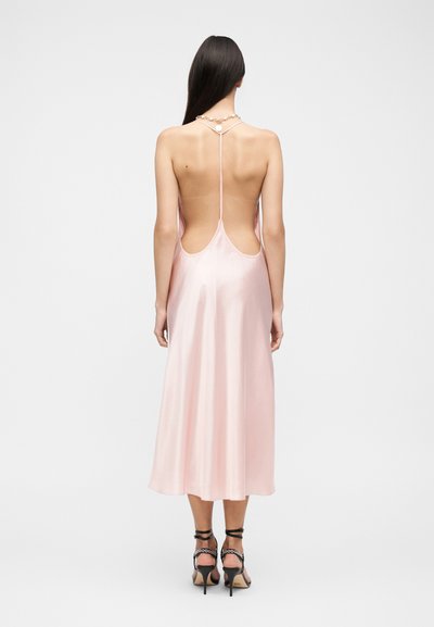 Γυναίκα με μακριά σκούρα μαλλιά που φοράει ένα ροζ σατέν μίντι φόρεμα χωρίς πλάτη και μαύρα ψηλοτάκουνα σανδάλια με λουράκια στον αστράγαλο.