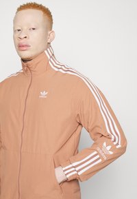 Gaiši brūns Adidas džemperis ar baltu trīs joslu dizainu uz piedurknēm, augstu apkakli un izšūtu logotipu uz krūtīm un piedurknēm.