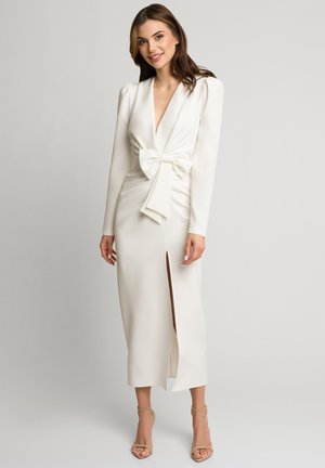 Kobieta ubrana w długą białą suknię z głębokim dekoltem w serek, bufkami na ramionach, dużą kokardą z przodu, rozcięciem na udzie oraz beżowymi szpilkami z paskiem wokół kostki.