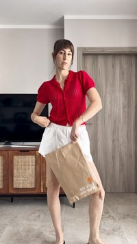 Cardigan corto a maniche corte di colore rosso, con colletto rotondo, abbinato a pantaloni bianchi. Tiene in mano una busta di carta marrone. Sullo sfondo, si vede un armadio di legno e una TV.