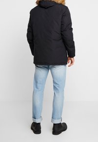 Sort dunjakke med pelsforet hætte, med lynlåslukning, sidelommer og en afslappet pasform. Kombineret med lyseblå op rullede jeans og sorte støvler.