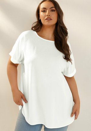 Frau mit langen dunklen Haaren, die ein locker sitzendes weißes Kurzarmoberteil und hellblaue Jeans trägt und vor einer schlichten Wand steht.