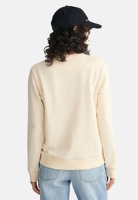 Cream-colored sweatshirt with a round neckline and long sleeves, soft texture, paired with light blue jeans and a black cap.