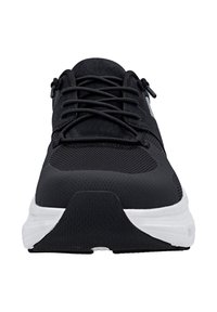 Scarpa sportiva nera con parte anteriore in rete, lacci incrociati e suola bianca spessa vista frontalmente.