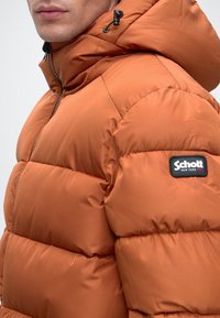Homme portant une doudoune orange avec capuche et un écusson logo Schott New York sur le haut du bras, légèrement de profil.