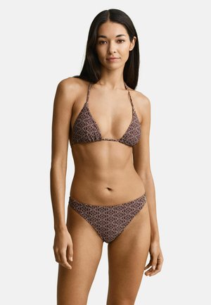 Jeune femme aux longs cheveux foncés portant un bikini marron avec des motifs géométriques clairs, se tenant devant un fond clair uni.