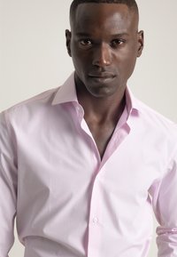 Camisa de vestir a rayas rosa claro con cuello abotonado, que presenta líneas verticales sutiles, textura suave y un diseño ajustado.