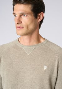 Maglione lavorato a maglia beige con scollo rotondo, caratterizzato da un bordo e polsini a costine, e un piccolo emblema bianco sul lato sinistro del petto. Tessuto leggero.