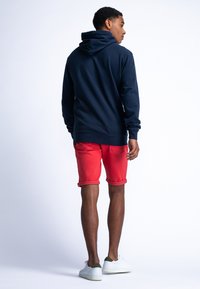 Marine hoodie met een capuchon, gecombineerd met rode shorts. De shorts zijn omgeslagen aan de zoom en witte sneakers maken de outfit compleet.