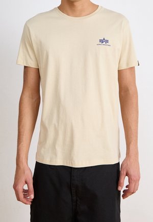 T-shirt beige à col rond en coton, avec un petit logo bleu "Alpha Industries" sur la poitrine et des manches courtes.