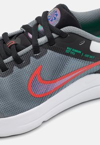 Baskets de sport grises avec une tige en maille texturée, des accents noirs, un logo Nike rouge vif et des détails de marque visibles. Semelle blanche en caoutchouc.