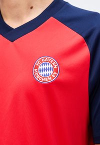 Κόκκινη και ναυτική φανέλα ποδοσφαίρου της FC Bayern München που φοριέται στον κορμό, με το έμβλημα του συλλόγου αριστερά στο στήθος.