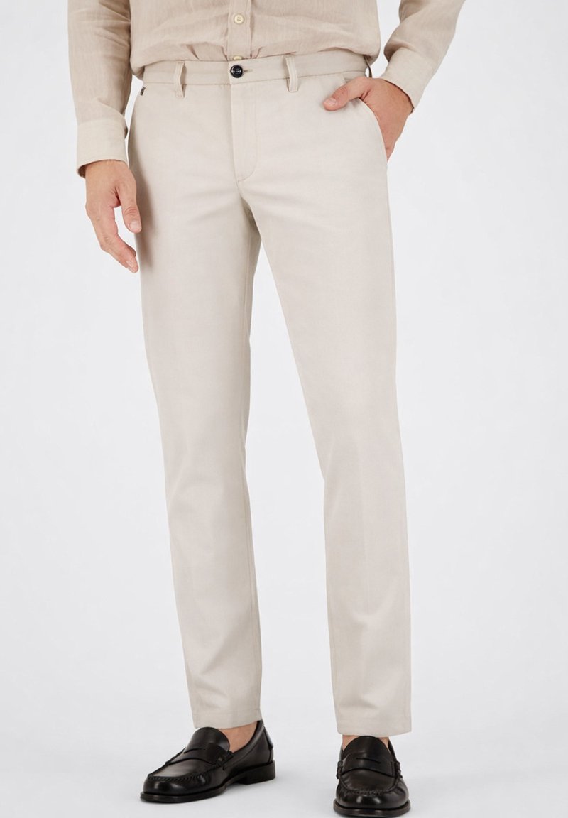 Homme portant un pantalon chino beige avec des mocassins noirs, main dans la poche, et une chemise beige boutonnée rentrée, debout devant un fond uni.