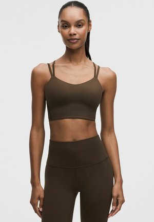 Donna con capelli scuri raccolti in una coda di cavallo che indossa un reggiseno sportivo aderente di colore oliva scuro con doppie spalline e leggings coordinati a vita alta.