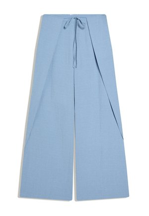 Młoda kobieta stoi z rękami w kieszeniach, ubrana w jasnoniebieską kamizelkę bez rękawów i spodnie o szerokich nogawkach, na tle omszałej kamiennej ściany i metalowego ogrodzenia.