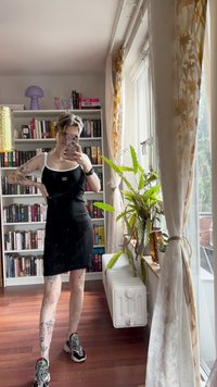 Juodas be rankovių suknelė su balta apdaila, glaudžiai priglundanti prie kūno, eksponuojama prieš knygų lentynų ir augalų foną, turinti tatuiruotes ant rankų.
