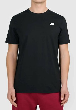 Mężczyzna noszący prostą czarną koszulkę z krótkim rękawem z małym białym logo "4F" na lewej piersi, w połączeniu z czerwonymi spodniami.