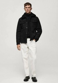 Giacca in shearling nera con chiusura a bottoni e tasche sul petto, indossata sopra un dolcevita nero e pantaloni bianchi, abbinata a calzature nere.