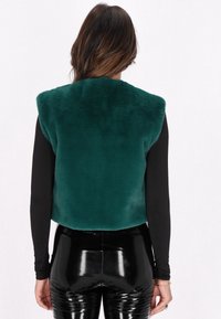 Vesta din blană sintetică într-o nuanță adâncă de teal, cu decolteu rotund, textură groasă și design fără mâneci, purtată peste colanți negri, lucioși.