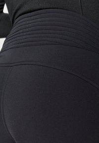 Visoko pasovi črne leggins hlače, izdelane iz raztegljive tkanine, z ribasto teksturo v predelu pasu in gladko površino na nogah.