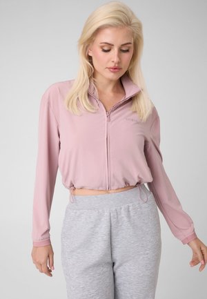 Blonde Frau in einer hellrosa kurzen Jacke mit Reißverschluss und hellgrauen Jogginghosen, die vor einem schlichten Hintergrund steht.