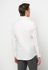 Camicia bianca a maniche lunghe realizzata in tessuto liscio, con colletto classico e polsini abbottonati, vista da dietro. Design semplice e minimale.
