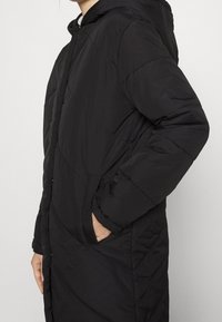Black, long padded coat with a hood, featuring a quilted design, side pockets, and concealed front closure. Smooth texture and lightweight.