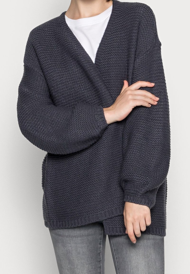 Personne portant un cardigan en maille texturée gris foncé sur une chemise blanche et un jean gris clair, les bras croisés devant.