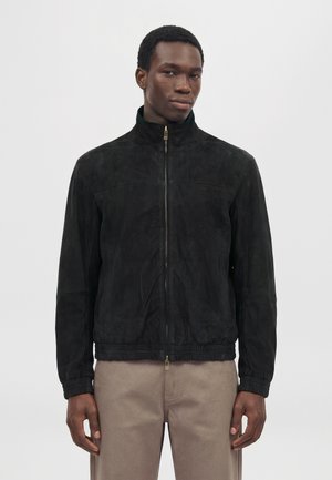 Un homme présente une veste noire zippée à col montant et un pantalon beige sur un fond clair uni.