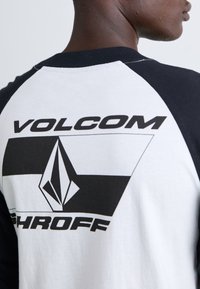 Hvid og sort raglan t-shirt med et stort sort grafisk print med "VOLCOM" og et diamantlogo på ryggen. Struktureret stof.